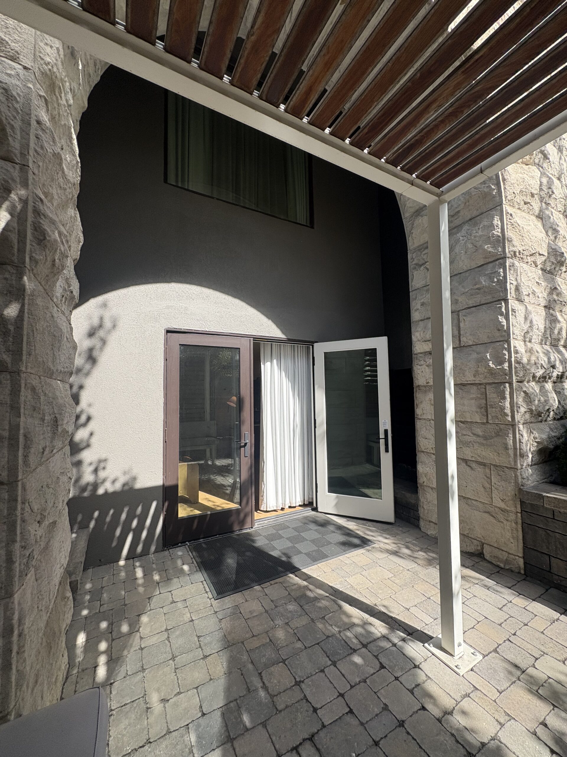 French doors of a hotel room opening onto a paver patio beneath a wood-slat pergola and stone arch