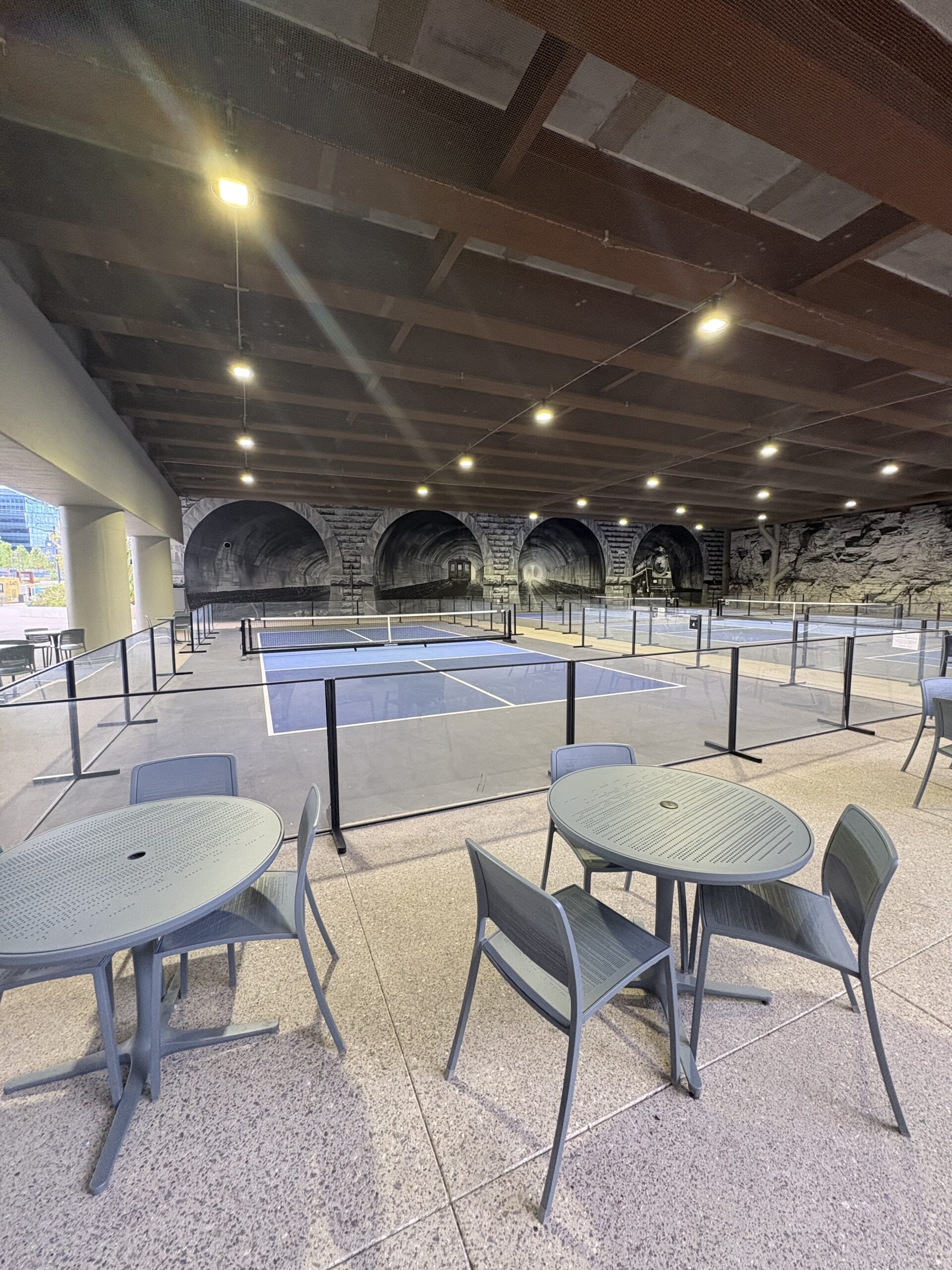 Covered pickleball courts under the Broadway Bridge with a vintage train tunnel mural on the back wall