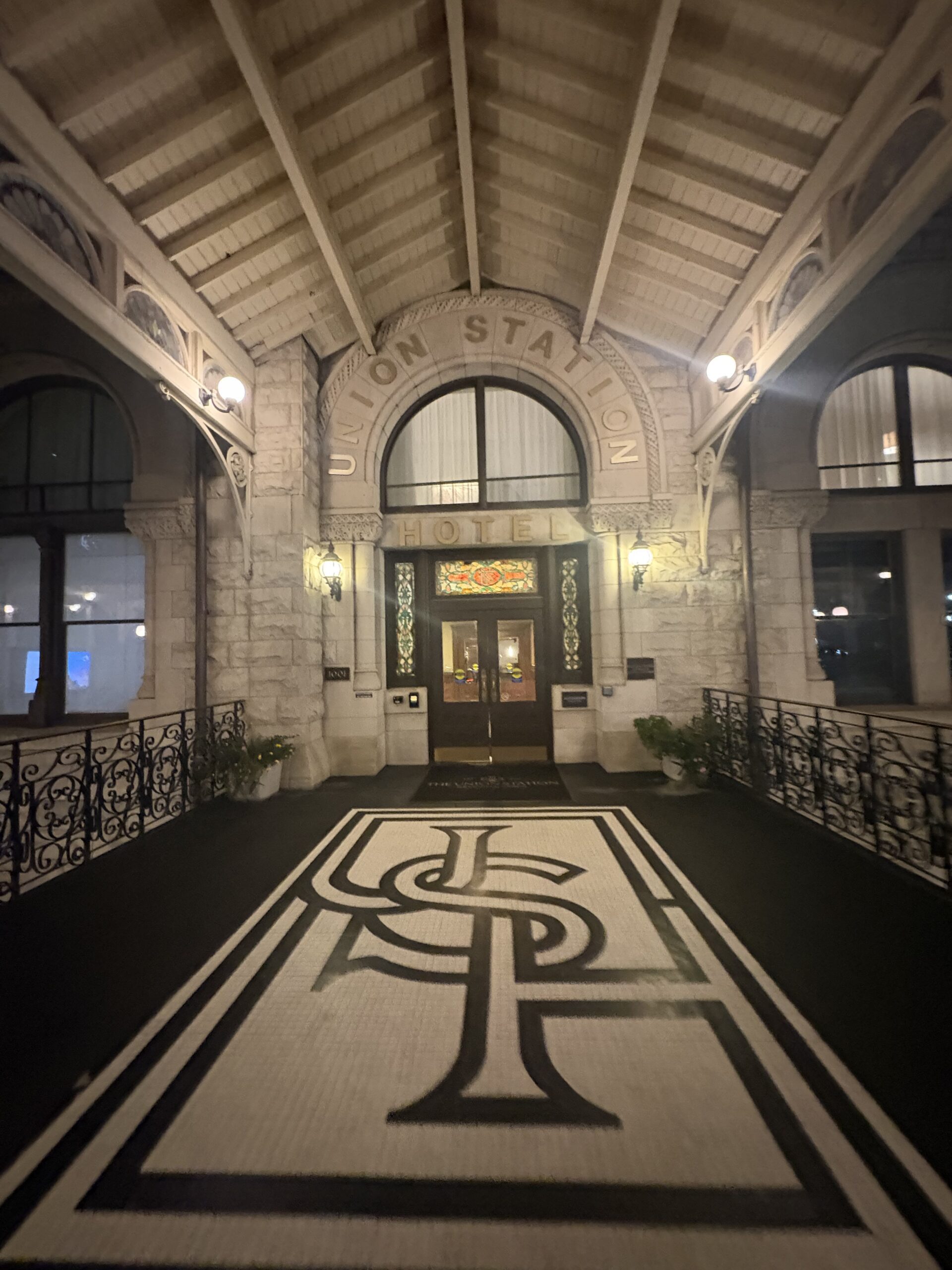 Union Station Hotel entrance at night with carved stone arch, stained-glass transom, and black-and-white monogram mosaic