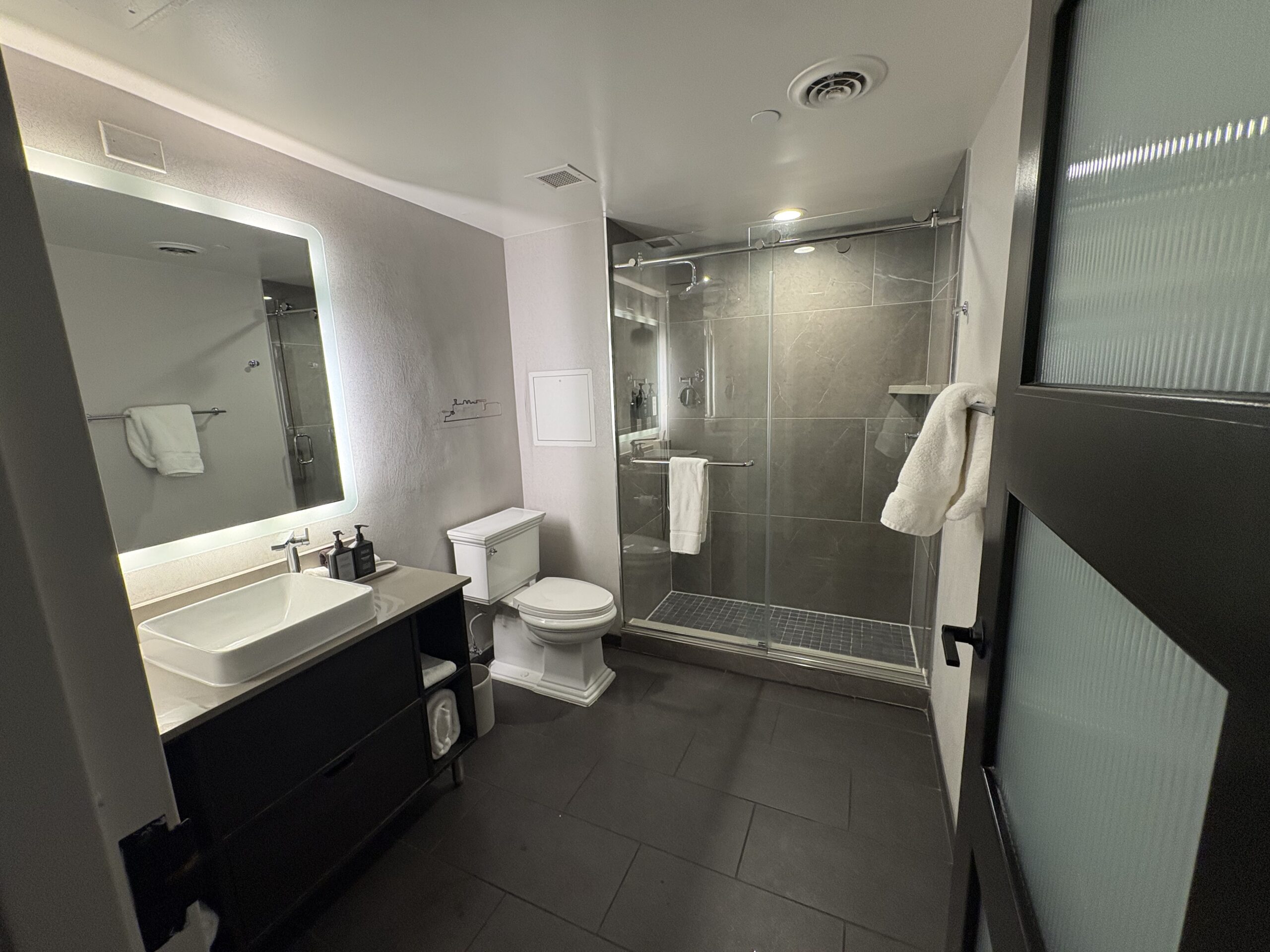 Hotel bathroom with backlit mirror, vessel sink, open-shelf vanity, and walk-in glass shower with rain showerhead
