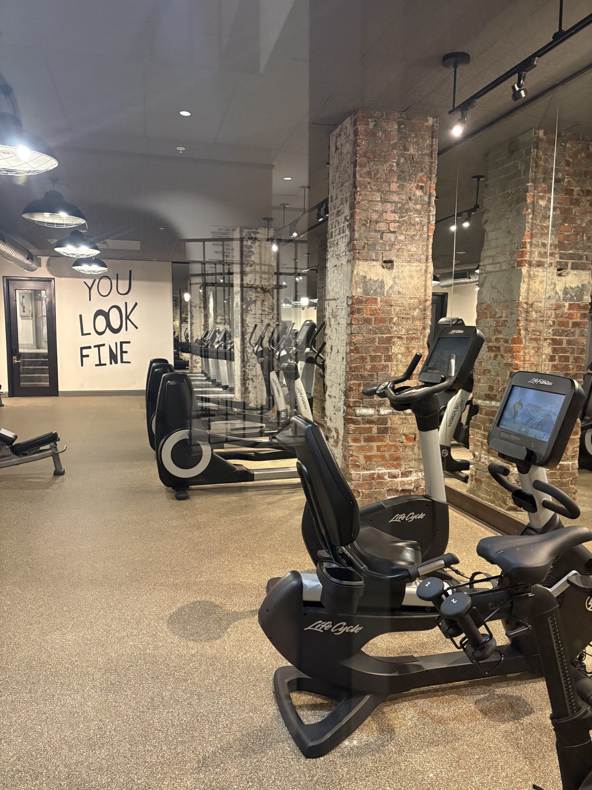 Hotel fitness center with Life Fitness bikes and ellipticals, exposed original brick columns, and a You Look Fine wall mural