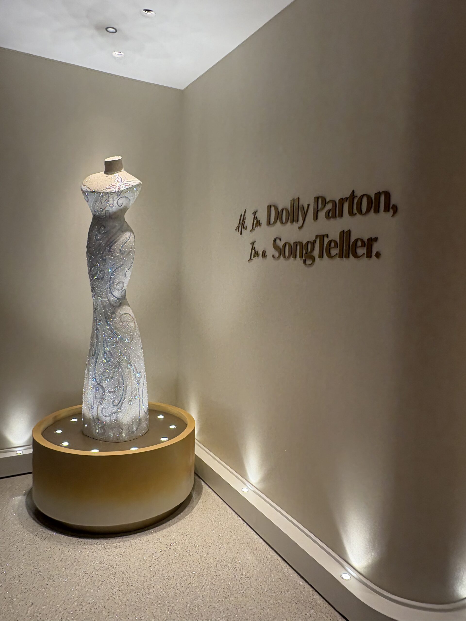 Crystal-embellished gown on a lit gold pedestal beside the SongTeller wall greeting in the hotel lobby